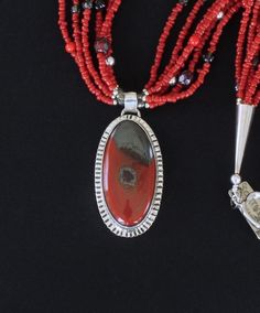 This appealing Necklace showcases a Rainbow Brecciated Jasper and Sterling Silver Pendant by Nevada Silversmith Jim Daggett. The Oval Pendant Stone is a vibrant brick red with black markings. It is wrapped in a raised Sterling Silver Bezel and set on Sterling Sheet cut to mirror the shape of the Stone. The Sheet has linear stamping that is radiant in reflected light. The Pendant drapes from a small Sterling Silver Cylinder Bail and is stamped on the back with the Artist's hallmark and "Sterling. Red Beaded Pendant Necklace With Natural Stones, Artisan Red Necklaces With Polished Beads, Artisan Red Necklace With Polished Beads, Red Gemstone Beads And Cabochons For Gifts, Artisan Red Jewelry With Gemstone Beads, Elegant Red Gemstone Beads And Cabochons, Unique Red Oval Beads Necklace, Unique Hand-strung Red Jewelry, Unique Red Oval Beaded Necklaces