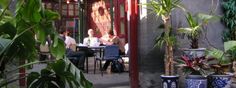 people are sitting at tables in an outdoor cafe with potted plants and large windows