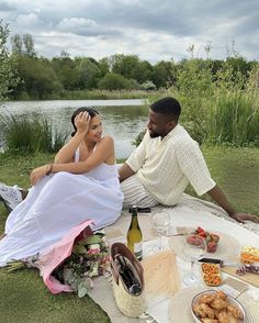 a man and woman sitting on the grass next to a body of water with food in front of them