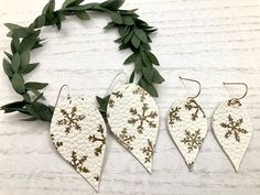 three white and gold leaf shaped earrings with green leaves around them on a wooden surface