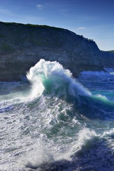 a large wave crashing into the ocean on a sunny day with mountains in the background