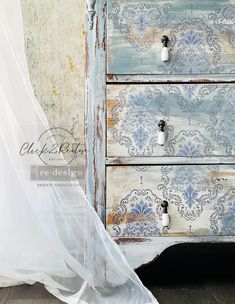 an old dresser painted in blue and white with flowers on the drawers, next to a sheer curtain