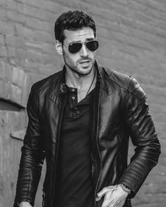 a man in black leather jacket and sunglasses standing next to a brick wall with his hands on his hips