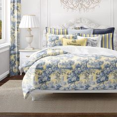 a white bed with blue and yellow comforter in a bedroom next to a window