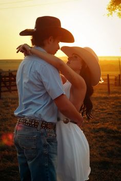 a man and woman standing next to each other in a field with the sun setting behind them