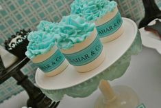 three cupcakes with blue frosting sitting on top of a white cake plate