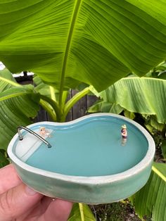 someone is holding a small blue bowl in front of some green plants and trees with their reflection on the water