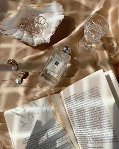 an open book sitting on top of a bed next to wine glasses and perfume bottles