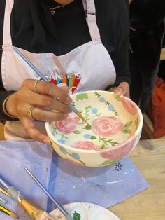 a woman in an apron painting a flowered bowl with paintbrushes on the table