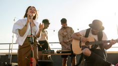 a woman singing into a microphone while playing the guitar on stage with other people standing around