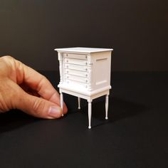 a hand is holding a miniature white dresser on a black surface with the drawer open