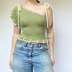 a woman wearing jeans and a green top with fringes on the shoulders is standing in front of a white wall