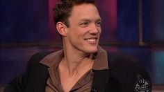 a man smiling while sitting down on a tv show in a brown shirt and black blazer