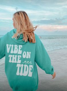 a woman is walking on the beach with her arms spread out to the side, wearing a blue hoodie that says vibe on the tide