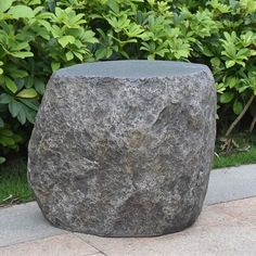 a large gray rock sitting on top of a sidewalk next to bushes and shrubbery