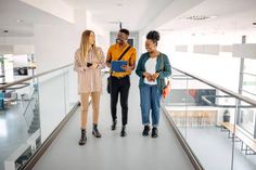 three young women standing on a walkway in an office building looking at something on the tablet