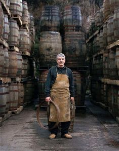 a man standing in front of barrels holding a large wooden barrel and wearing an apron