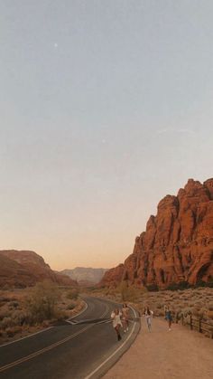 people are walking down the road in front of some red rocks and mountains at sunset