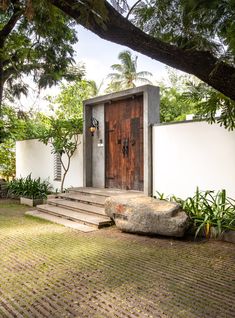 a house with a wooden door and steps leading up to the front door is surrounded by greenery