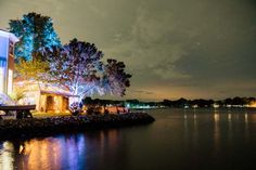 a house is lit up at night on the water's edge with trees and lights reflected in the water