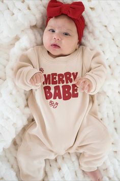 A baby in a beige holiday-themed onesie that reads "Merry Babe" lies on a textured white blanket. The baby wears a red headband with a big bow and looks up with wide eyes. Cozy Holiday, Holiday Ready, Holiday Memories, Holiday Gathering