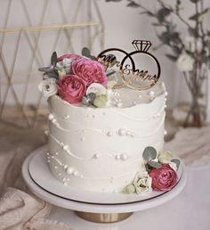 a white wedding cake with pink flowers and two rings on top, sitting on a table