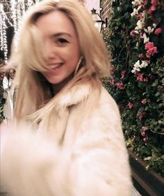 a woman in a fur coat is smiling at the camera while standing next to flowers