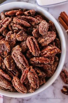 a white bowl filled with pecans next to cinnamon sticks on a marble counter top