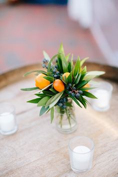 a vase filled with oranges and greenery on top of a wooden table next to candles