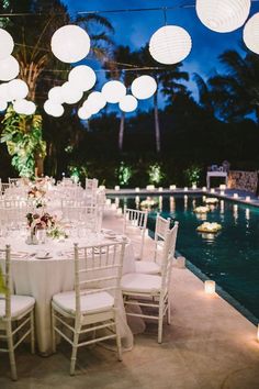 a long table with white chairs and paper lanterns hanging from it's ceiling next to a pool