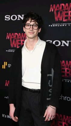 a man with glasses standing in front of a red and black sign that says madam west