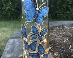 a glass vase with blue butterflies painted on it sitting on a stone slab in front of some trees