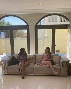 two women sitting on a couch in front of large windows