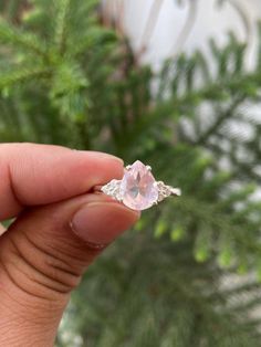 a person holding a ring with a pink stone in it's middle and green plants behind them