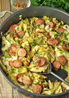 a pan filled with sausage and cabbage on top of a wooden table next to a spoon