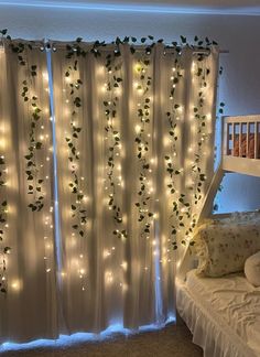 a white bed sitting next to a window covered in curtains and lights with green leaves on them