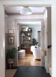 an open door leading to a living room and dining area with white furniture, potted plants and pictures on the wall