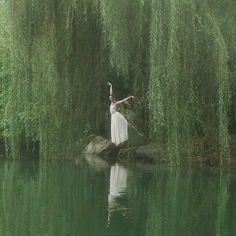 a woman in a white dress is standing on a rock by the water with willow branches