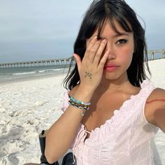 a woman in pink shirt holding her hand up to her face on beach with pier in background