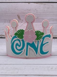 a pink and blue one birthday crown on top of a white wooden table with flowers