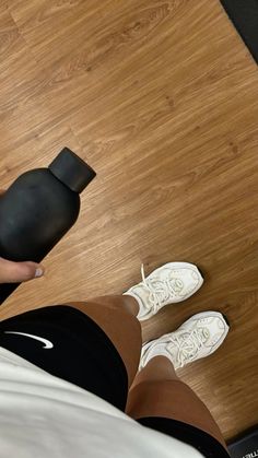 a person standing on a wooden floor holding a black object in one hand and white shoes on the other