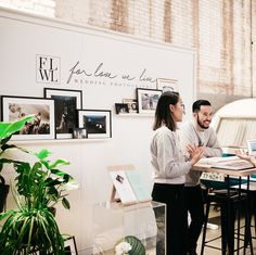 two people standing at a table talking to each other in front of pictures on the wall