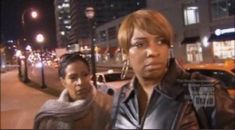 two women standing on the side of a street at night with buildings in the background