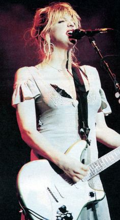 a woman holding a guitar while singing into a microphone and wearing a white dress with ruffled sleeves