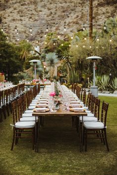 a long table is set up for an outdoor wedding reception with hanging lights and greenery