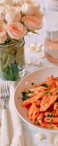 a white plate topped with pasta next to a vase filled with pink roses and water