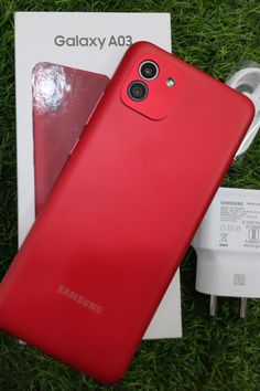 a red samsung phone sitting on top of a grass covered field next to a charger