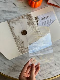 a person holding an open wedding card on top of a marble table with pumpkins