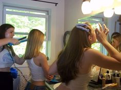 three young women blow drying their hair in front of a mirror while another woman looks on