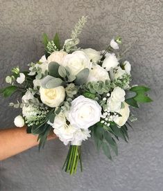 a bridal bouquet with white flowers and greenery on a wall in front of a person's hand